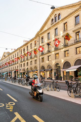 Fototapeta premium Geneva, Switzerland - August 30, 2016: People on motorcycle and bicycles on Rue de la Corraterie Street with Swiss flags in the city center of Geneva, Switzerland
