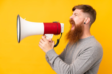 Redhead man with long beard over isolated yellow background shouting through a megaphone