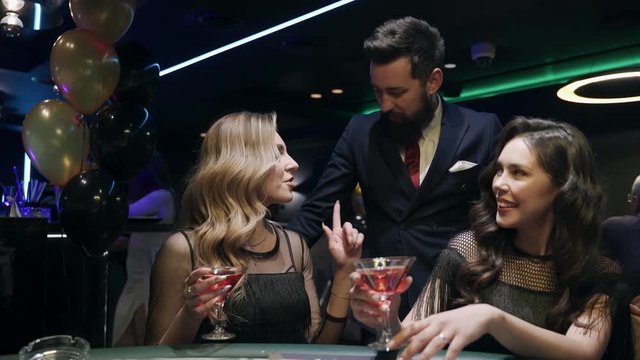 Casino: A bearded man communicates with preatty girls and they smile and laugh. Аlcoholic red champagne in the hands. They are sitting at the pocker table