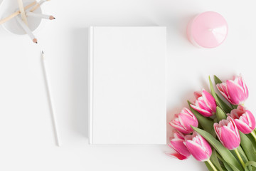 Top view of a book mockup with a bouquet of pink tulips and workspace accessories on a white table.