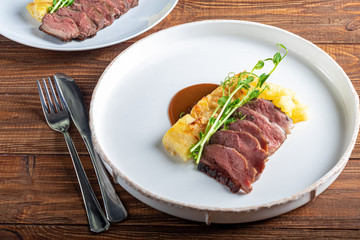 Duck breast with a side dish of potato casseroles, seasoned with pea microgreen with tomato and chili sauce. On a white plate. On a wooden background,