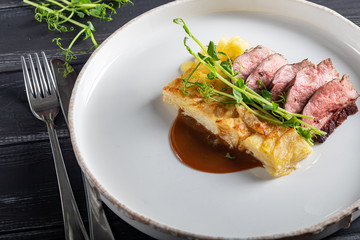 Duck breast with a side dish of potato casseroles, seasoned with pea microgreen with tomato and chili sauce. On a white plate. On a wooden background,