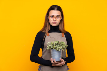 Young gardener girl holding a plant over isolated yellow background keeping arms crossed