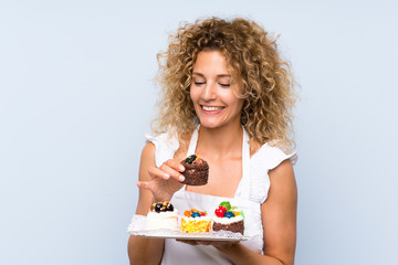 Young blonde woman with curly hair holding lots of different mini cakes