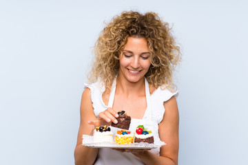 Young blonde woman with curly hair holding lots of different mini cakes