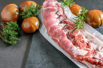 Big piece of fresh baby-back ribs for cooking, some fresh herbs and dark tomatoes on the black shale kitchen board.