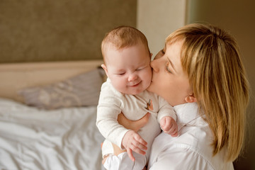 mother plays with a baby of 6 months of European appearance, both in white clothes are lying on the bed