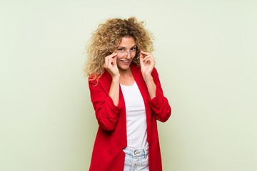 Young blonde woman with curly hair over isolated green background with glasses and surprised