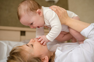 mother plays with a baby of 6 months of European appearance, both in white clothes are lying on the bed