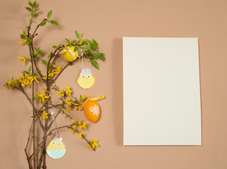 Easter composition. Paper blank, sprigs of forsythia, colored eggs and wooden Easter decorations on a beige background. Free space.