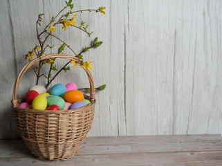 Easter composition. Basket with colorful eggs and spring branches of forsythia with leaves and yellow flowers on a wooden background. Free space.