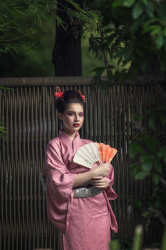 Woman dressed in a geisha costume, standing in the garden, holding a hand fan