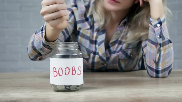 Young  woman putting a coins into glass jar with label of BOOBS text. Saving money concept