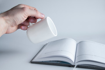 An open book or notepad with empty blank pages. White cup in hand pours on a book. On a white or gray background