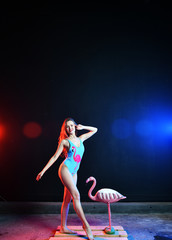 Fototapeta premium beautiful sexy girl with red hair in a swimsuit with a flamingo pattern posing with a pink flamingo toy in colored smoke