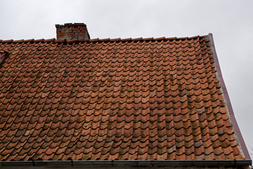 A fragment of an old shabby brown beige gable tiled roof of an old house with a brick chimney stack furnace with a rain gutter under it against the background of a gloomy gray overcast sky
