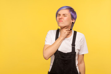 Flu, throat pain. Portrait of unhealthy girl with violet short hair in denim overalls clutching neck and frowning from ache, suffering sore throat, influenza. isolated on yellow background studio shot
