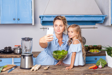 Charming woman and little girl taking selfie in modern kitchen, holding bowl of vegetable salad and...