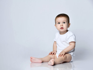 Infant baby boy toddler in white bodysuit is sitting sideways on the floor looking up at us on a gray with copy space