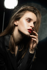 Beautiful braless slim girl, wearing a unbuttoned black blazer, with a slight frenzy and detachment in her eyes touches her face with her hand on a dark background, next to the light lamp.