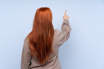 Redhead teenager girl with sweater over isolated blue background pointing back with the index finger