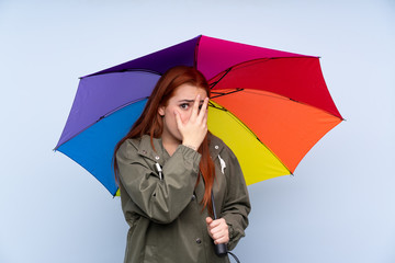 Redhead teenager girl holding an umbrella over isolated blue background covering eyes and looking through fingers