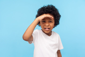 Portrait of astonished little boy with curls holding hand over eyes and looking attentively far...