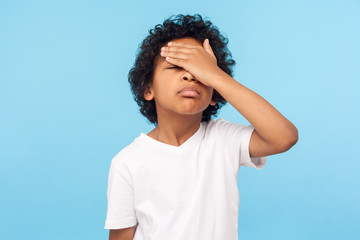 Fototapeta na wymiar Facepalm. Portrait of forgetful upset little boy with curls covering face with hand and expressing sorrow regret for mistake, forgot to do homework. indoor studio shot isolated on blue background