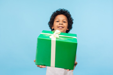 Take this present! Happy generous good-natured little boy with curly hair giving gift box and looking at camera with toothy smile, holiday charity. indoor studio shot isolated on blue background