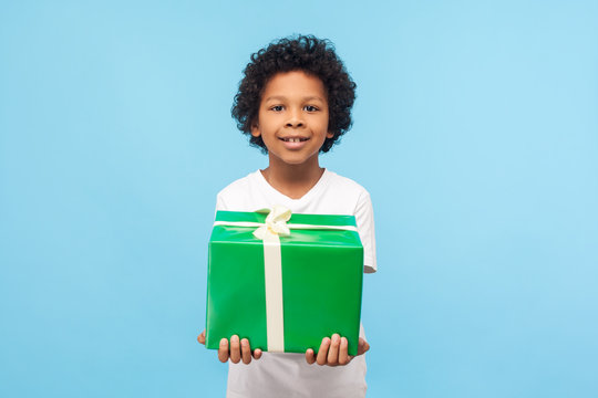 Child Birthday Celebration. Positive Cute Joyful Little Boy With Curly Hair Holding Large Wrapped Gift Tightly With Two Hands And Smiling Happily At Camera. Studio Shot Isolated On Blue Background