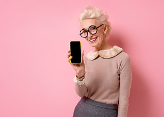 Smiling grandmother shows empty screen phone. Photo of kind elderly woman on pink background.