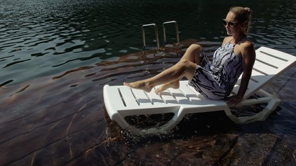 Woman lie on a sunbed in sunglasses and a boho silk shawl. Girl rest on a flood wood underwater pier. The pavement is covered with water in the lake. - obrazy, fototapety, plakaty