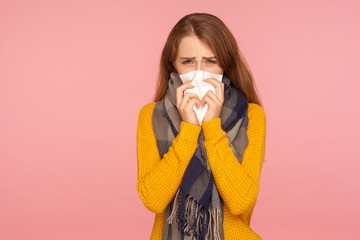 Flu season. Portrait of sick red hair girl wearing big scarf, sneezing and blowing nose, wiping with tissue, suffering temperature and cough, influenza. indoor studio shot isolated on pink background
