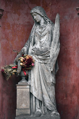 A weathered sandstone sculpture of a grieving angel with flowers in her hand in front of a red...