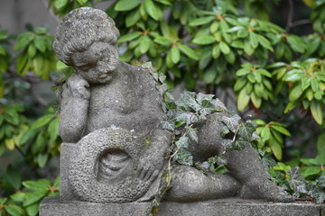 A weathered, nearly overgrown  sandstone sculpture of a resting angel on a tomb of a cemetery in Berlin-Germany.