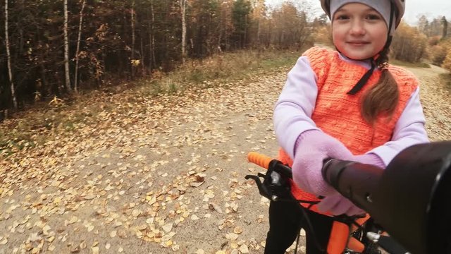 Take selfie on camera phone. One caucasian children rides bike road in autumn park. Girl riding black orange cycle in forest. Kid goes do bicycle sports. Biker motion ride with backpack and helmet.