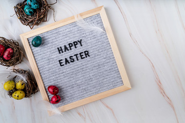 The words Happy Easter on grey felt letter board decorated with quail and chicken eggs and feathers on marble background