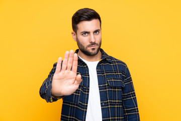 Young handsome man with beard over isolated yellow background making stop gesture