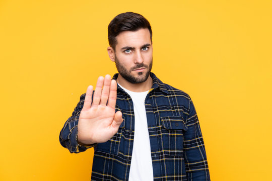 Young handsome man with beard over isolated yellow background making stop gesture