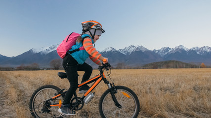 Fototapeta premium One caucasian children rides bike in wheat field. Little girl riding black orange cycle on background of beautiful snowy mountains. Biker motion ride with backpack and helmet. Mountain bike hardtail.