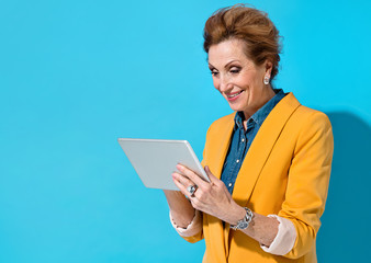 Smiling elderly woman using digital tablet. Photo of business lady in yellow blazer on blue background. Emotions and pleasant feelings concept.