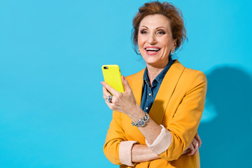 Happy elderly woman holds phone. Photo of business lady in yellow blazer on blue background. Emotions and pleasant feelings concept.