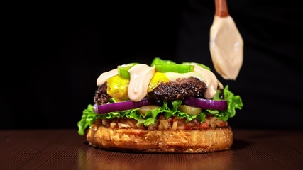 Craft burger is cooking on black background. Consist: sauce salsa, lettuce, red onion, pickle, cheese, chilli green pepper, bun, marbled meat beef.