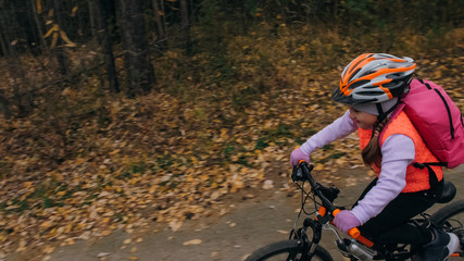 Fototapeta premium One caucasian children rides bike road in autumn park. Little girl riding black orange cycle in forest. Kid goes do bicycle sports. Biker motion ride with backpack and helmet. Mountain bike hard tail.