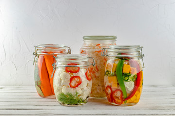 glass jars of fermented cabbage, cauliflower, cucumbers, onions, carrots, chili pepper, vegetables on a light background. fermentation is a source of probiotics.