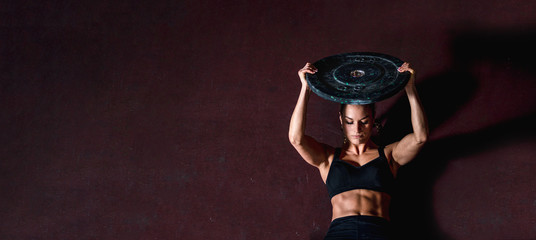 Young strong sweaty fit muscular girl with big muscles doing sit ups with barbell weight plate for abdominal muscles or abs hard crossfit workout training on the gym floor view from above real people