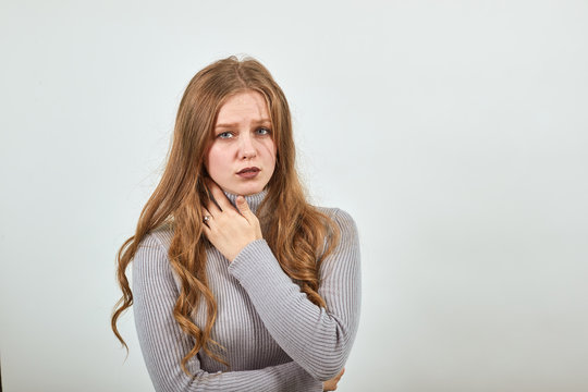 A young beautiful woman in gray sweater with red hair is bent in pain, holding her neck and throat sore