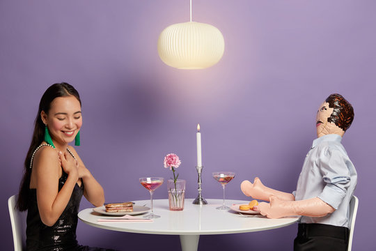 Touched woman pretends receiving heartwarming compliment from lover, sits at table with layman doll, keeps hands on chest, smiles pleasantly during romantic date, pose at cozy restaurant, have dinner
