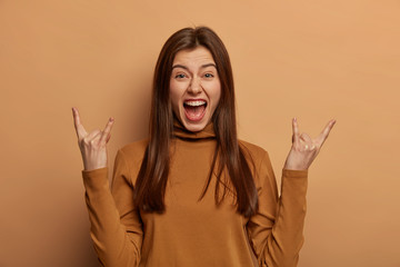 Emotional womann shows horns up gesture, returned from rock n roll concert, likes awesome music, exclaims loudly, wears brown turtleneck, isolated on beige studio wall, feels happy, free and wild.