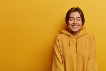 Fototapeta premium Lifestyle and emotions concept. Glad woman closes eyes and chuckles, laughs and has fun, wears sweatshirt, shows white teeth, stands against yellow background, blank space for your advertisement.